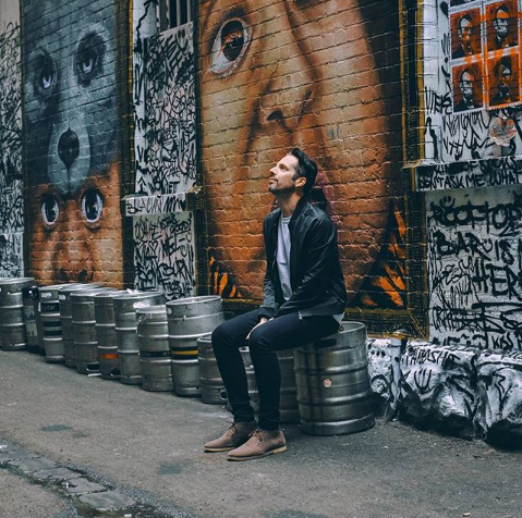 Man sitting on a keg, looking up at graffiti-covered brick wall with large eyes mural.
