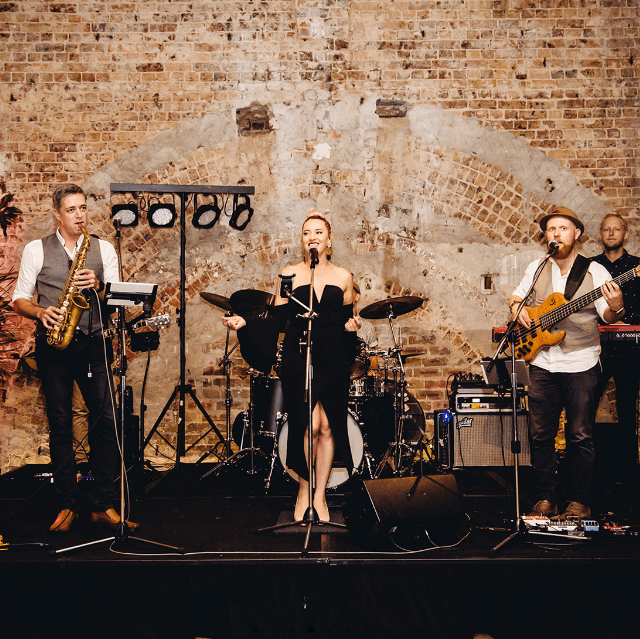 A band performs on a stage against a textured brick wall, featuring a singer, a saxophonist, a bassist, and a drummer.