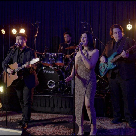 A four-piece band performs on stage with a singer, guitarist, bassist, and drummer in a dimly lit, professional setting.