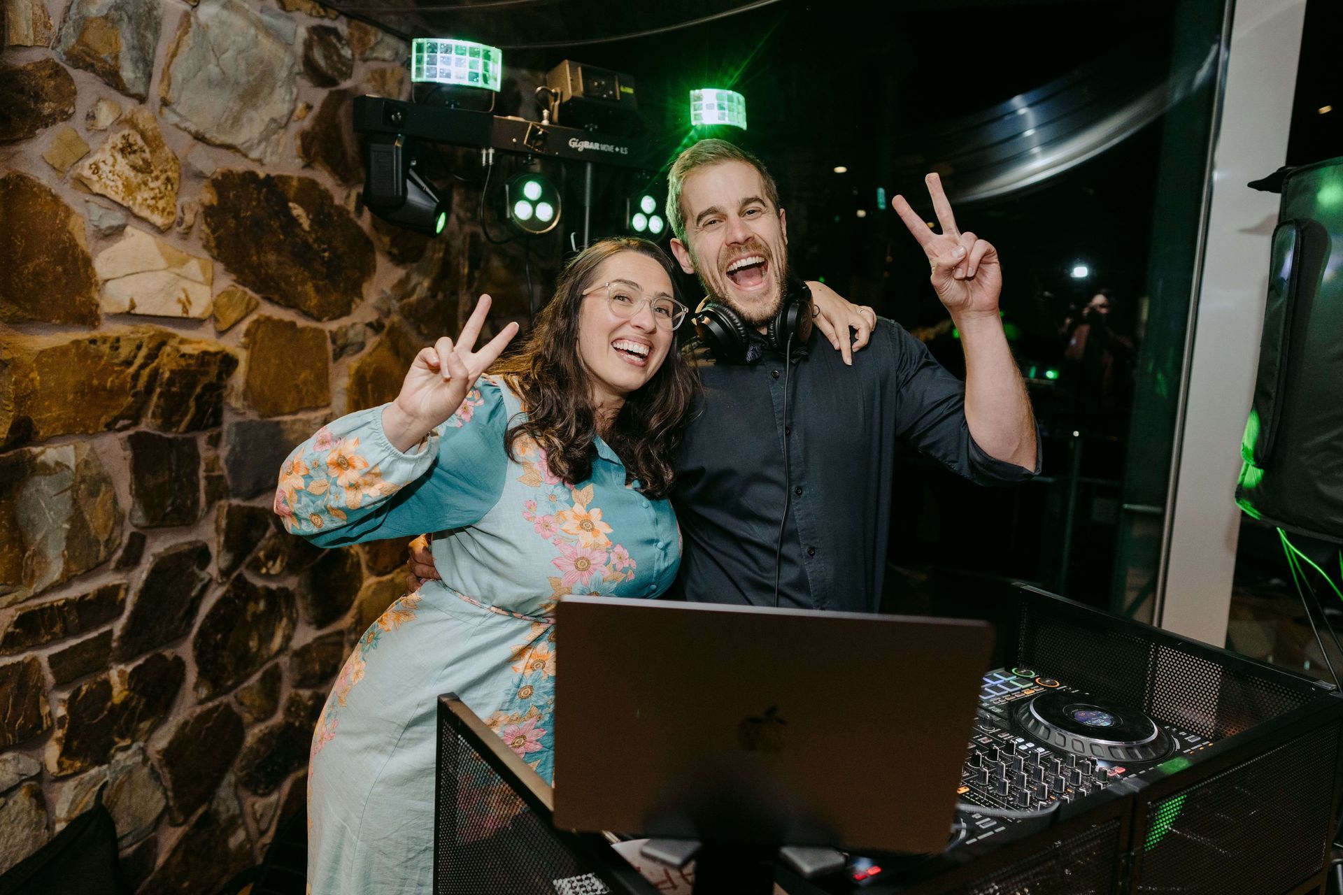 Woman and DJ with peace signs, smiling, by a laptop and music gear. Stone wall in background.