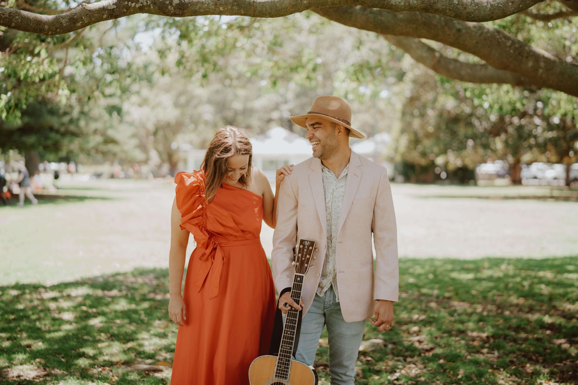 A man and a woman are standing under a tree holding hands and a guitar.
