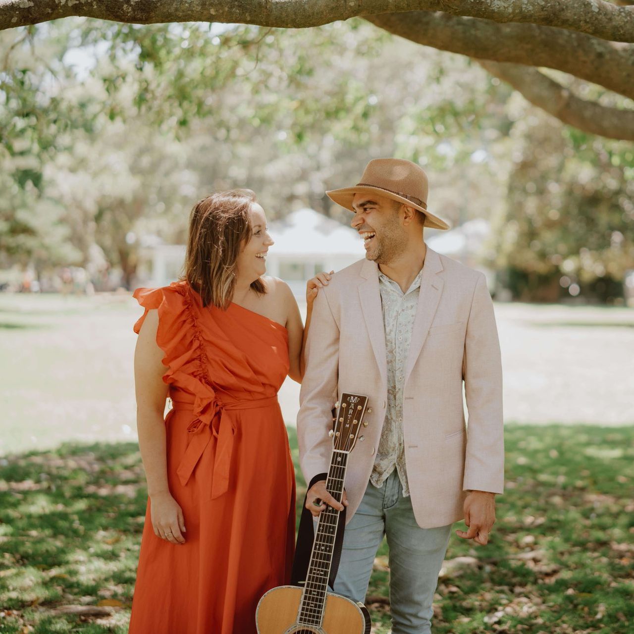 A person in an orange dress and another in a blazer and hat stand outdoors, laughing and holding an acoustic guitar.