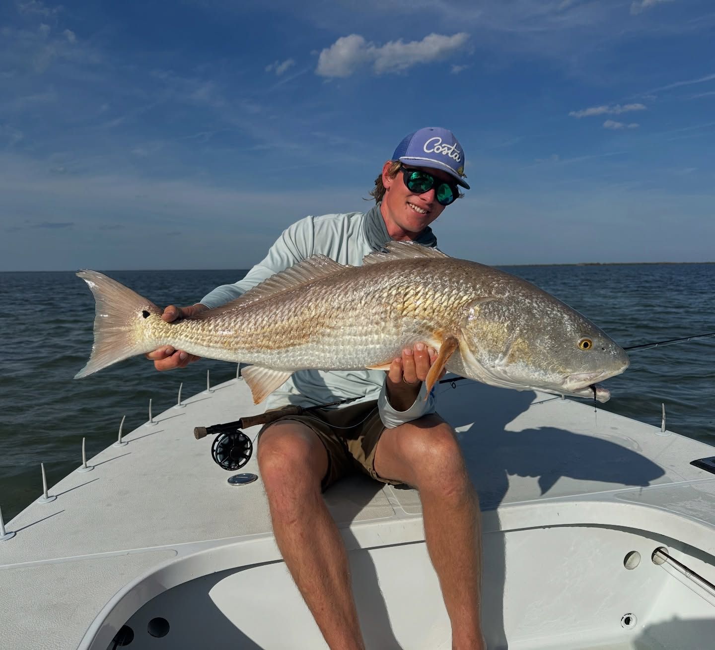 Person on a boat holding a large redfish, with blue sky and open water behind.