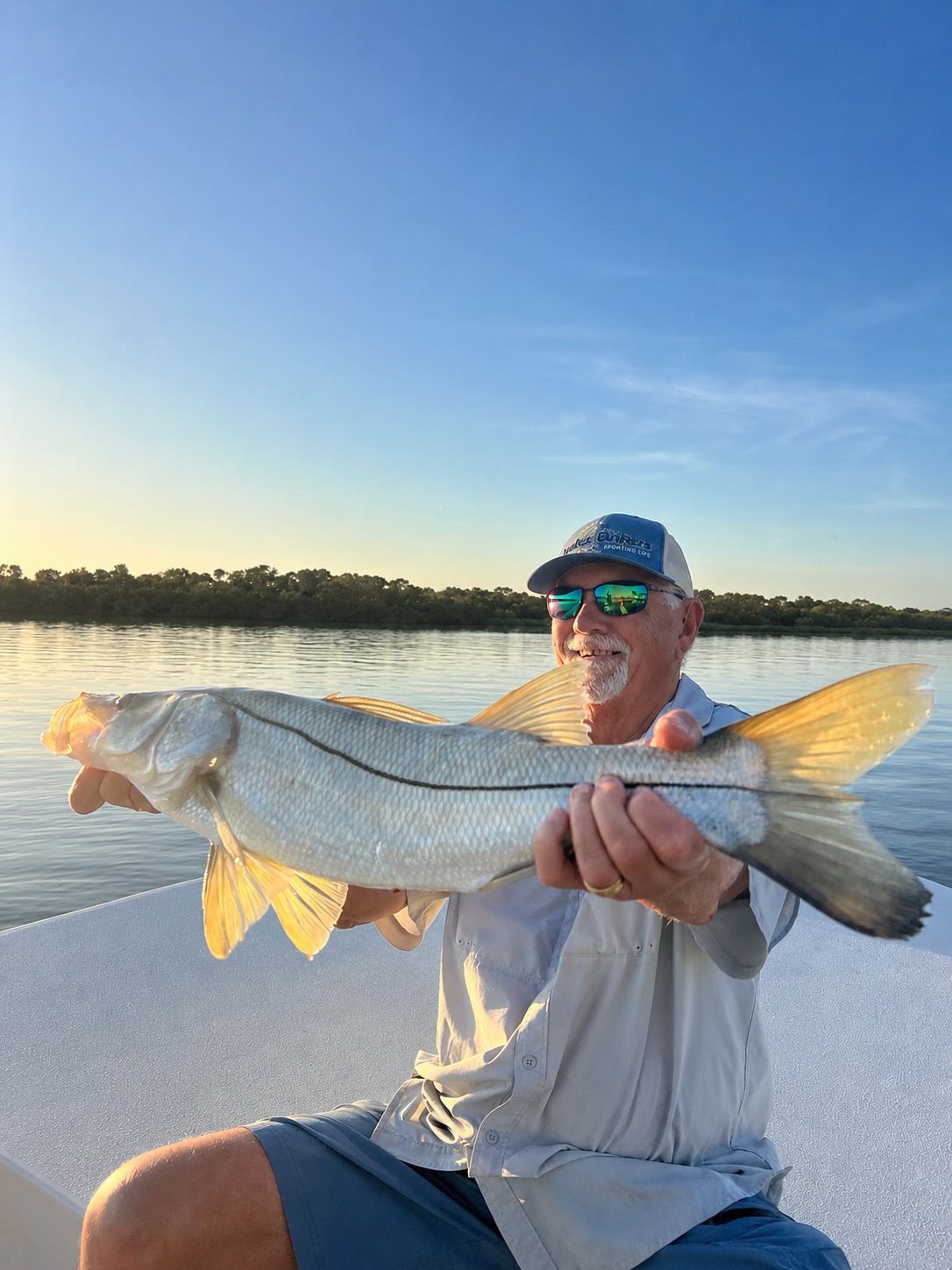 Man holding a large silver fish at sunset on a boat, with calm water and shoreline in the background