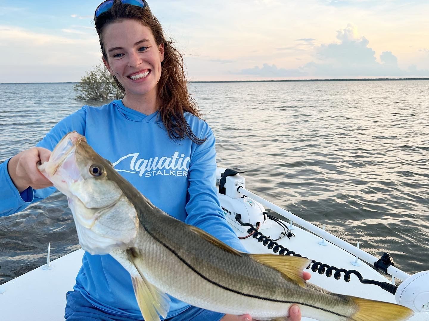 Smiling person on a boat holding a large striped fish at sunset on calm water