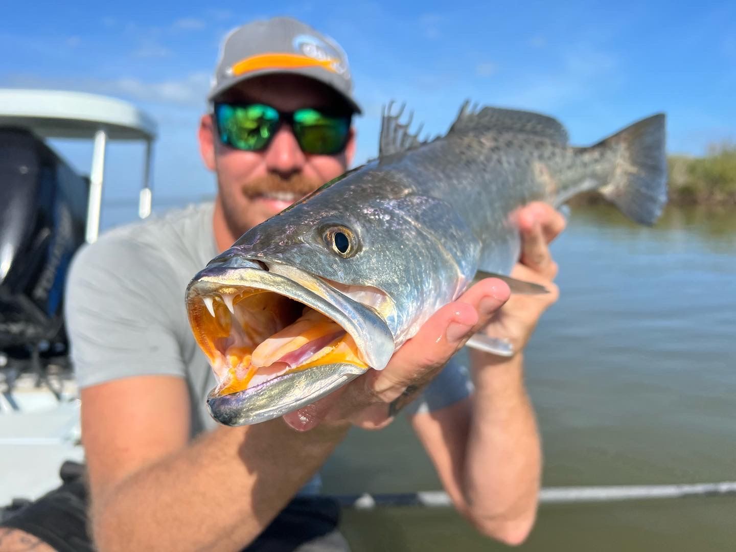 Person holding a large fish on a boat, wearing sunglasses and a cap, with the fish’s mouth open.