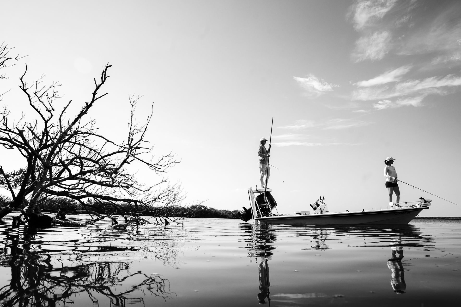 Two people stand on a boat in calm water beside a leafless tree, with reflections in black and white.