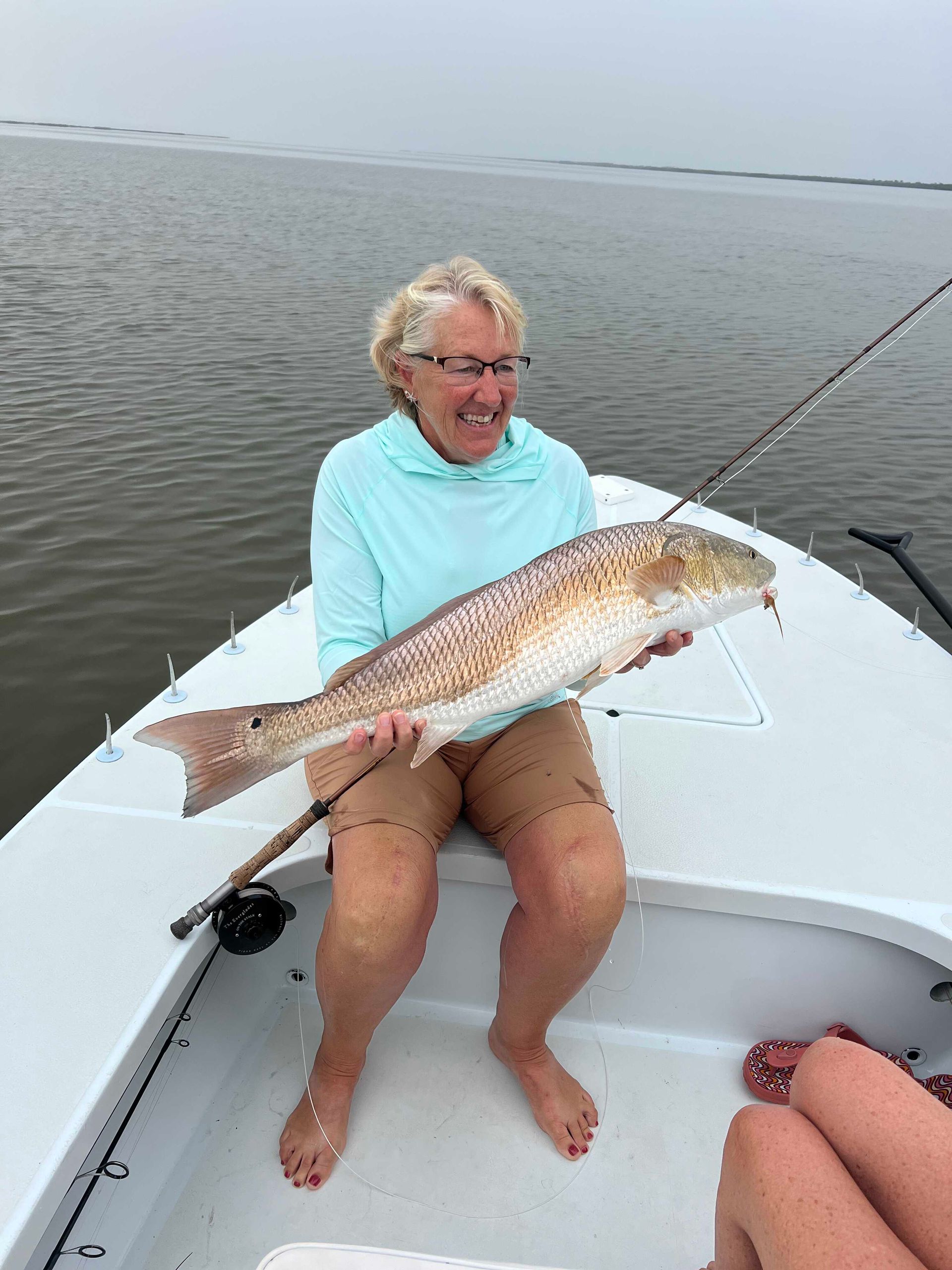 Woman on a boat holding a large fish, smiling on calm water.