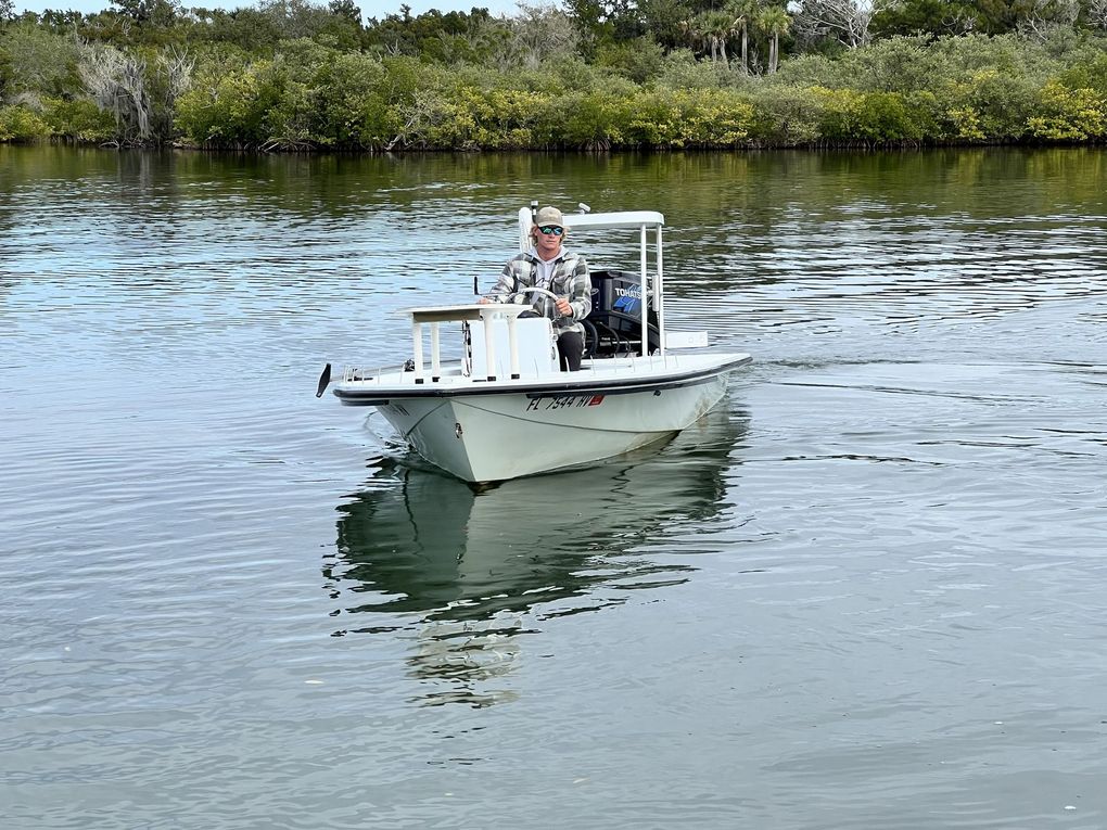 Small white motorboat on calm water with two people, near mangroves and shoreline.