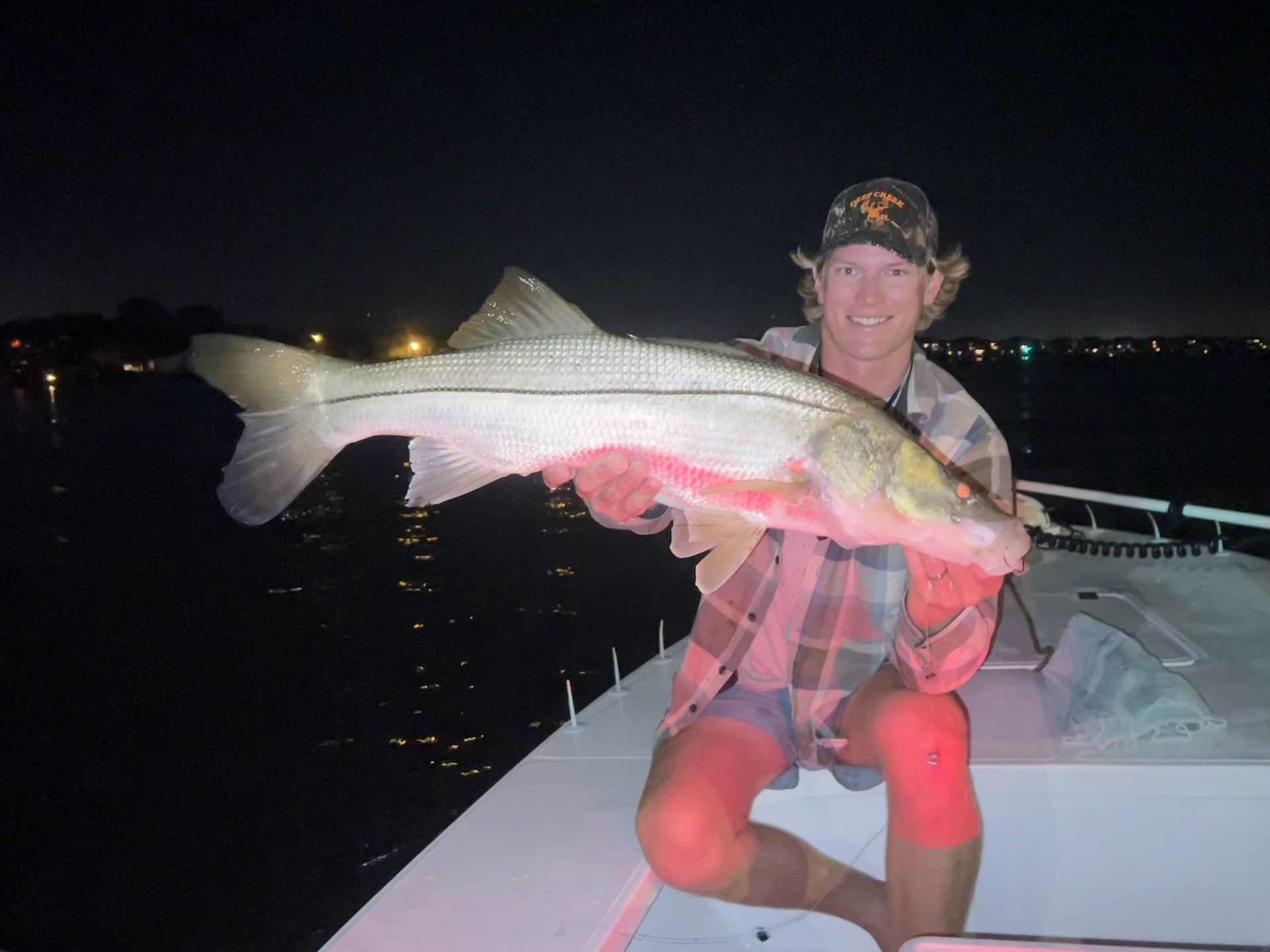 Man holding a large striped fish on a boat at night, with city lights in the background