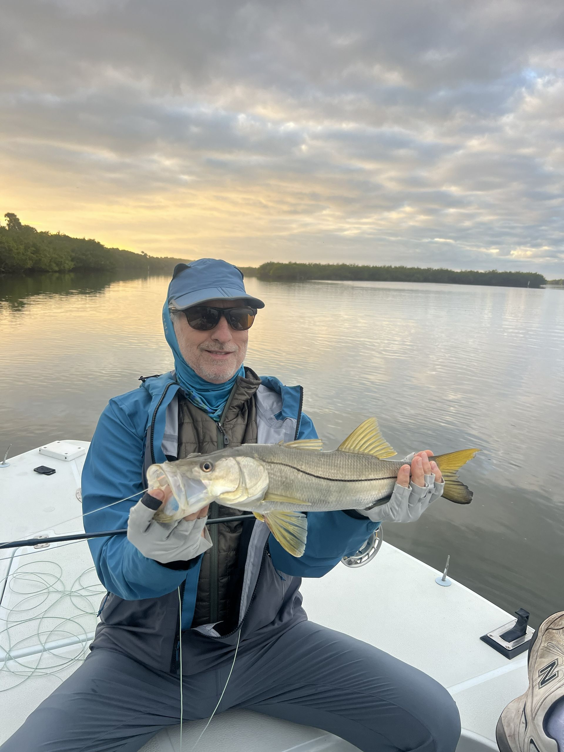 Angler in blue holds a large fish on a boat at sunset on a calm lake.