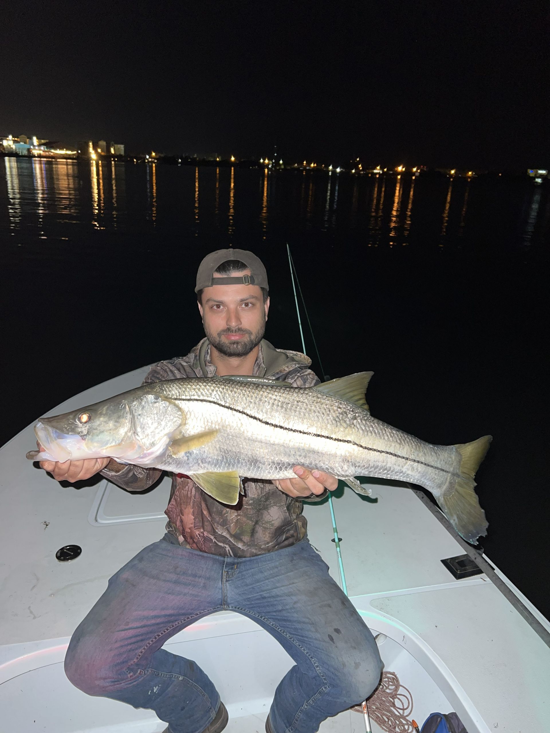 Man sitting on a boat at night, holding a large fish with city lights reflecting on the water.