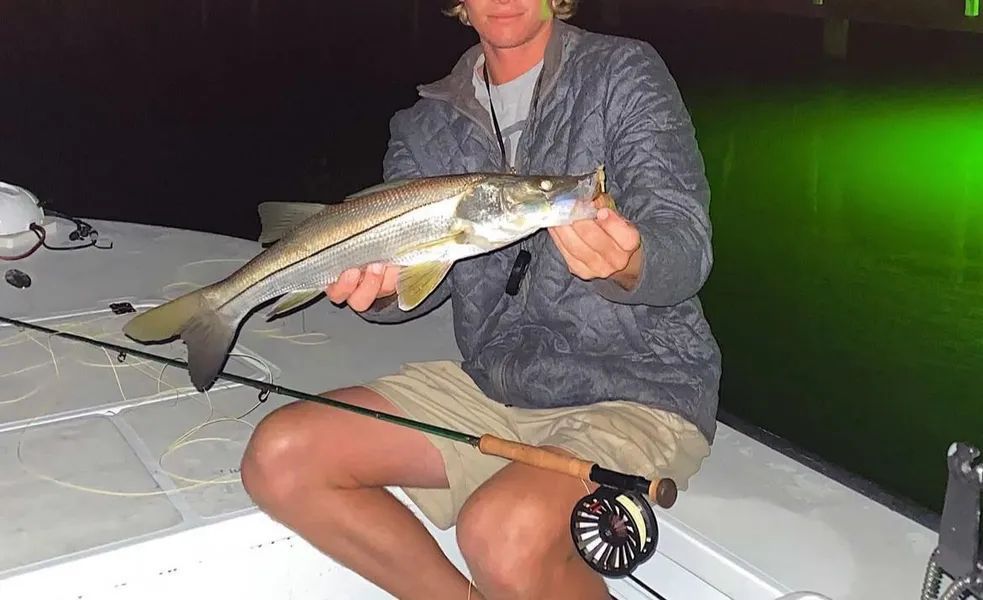 Person sitting on a boat at night, holding a large fish with green water glowing in the background.