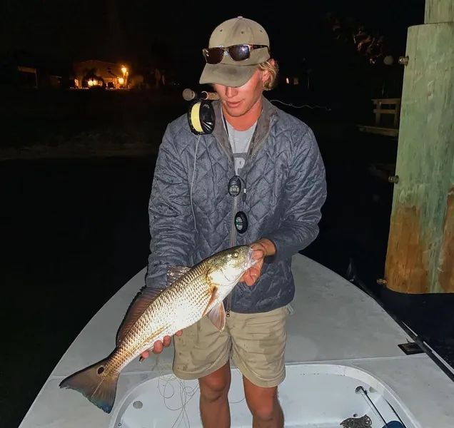 Person holding a large fish on a boat at night, wearing a cap and jacket.