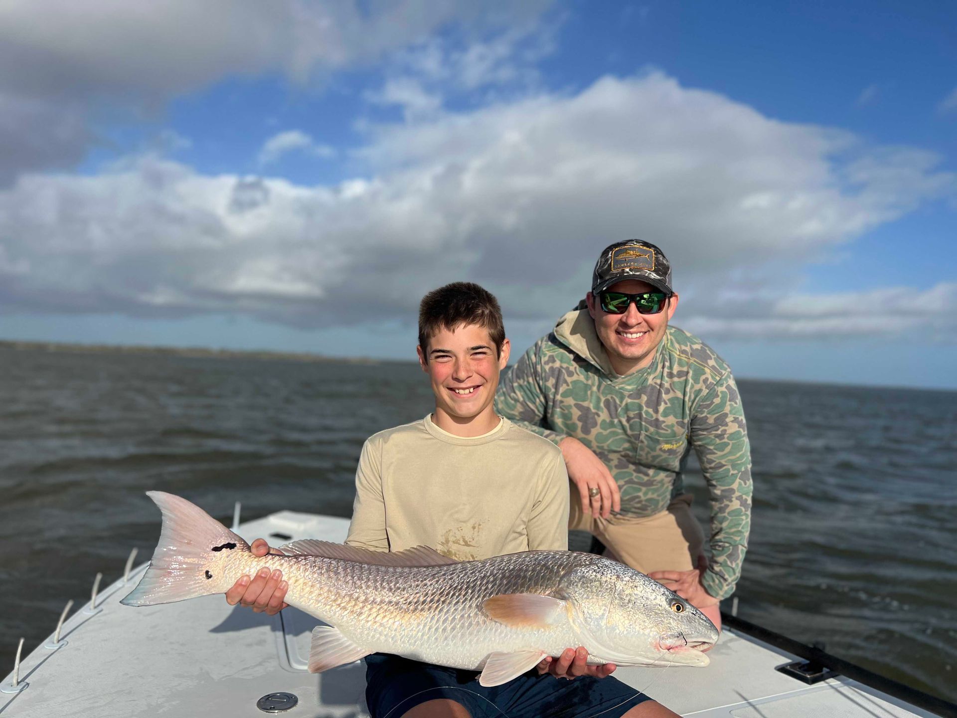 Two people on a boat holding a large fish with calm water and cloudy sky in the background
