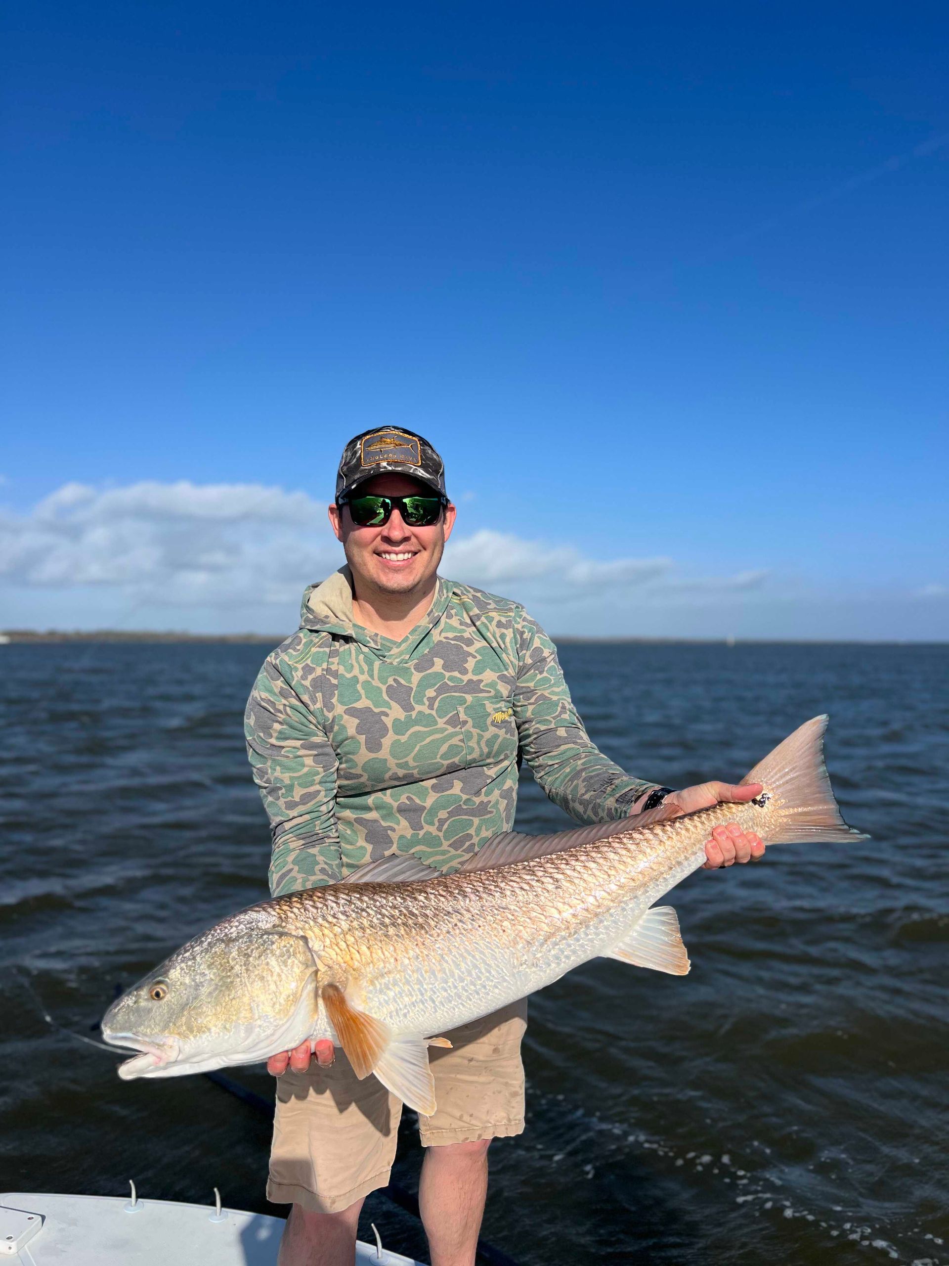 Smiling person in camo shirt holding a large fish on a boat with open water behind them