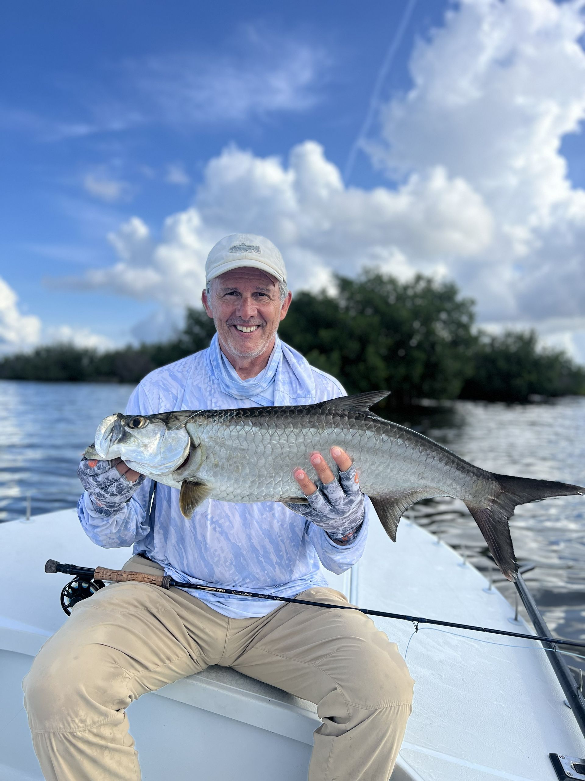 Man holding a large fish on a boat, smiling on calm water under a cloudy sky.