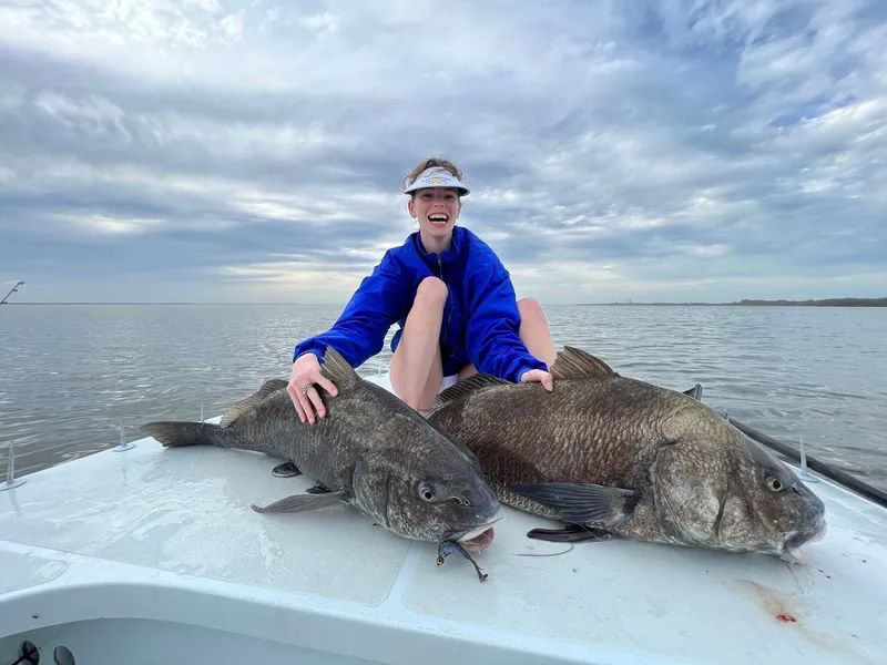 Person in blue jacket sits on a boat with two large fish on the deck under a cloudy sky