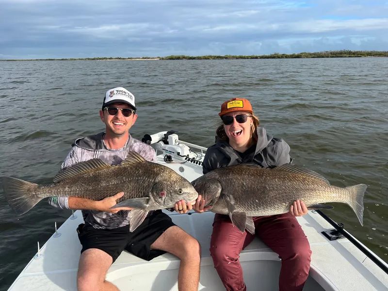 Two people on a boat holding large fish on a calm lake under a cloudy sky