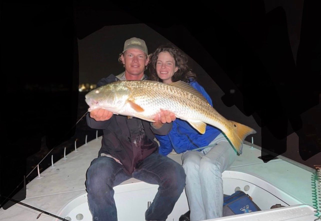 Two people on a boat at night holding a large fish, with city lights in the background.