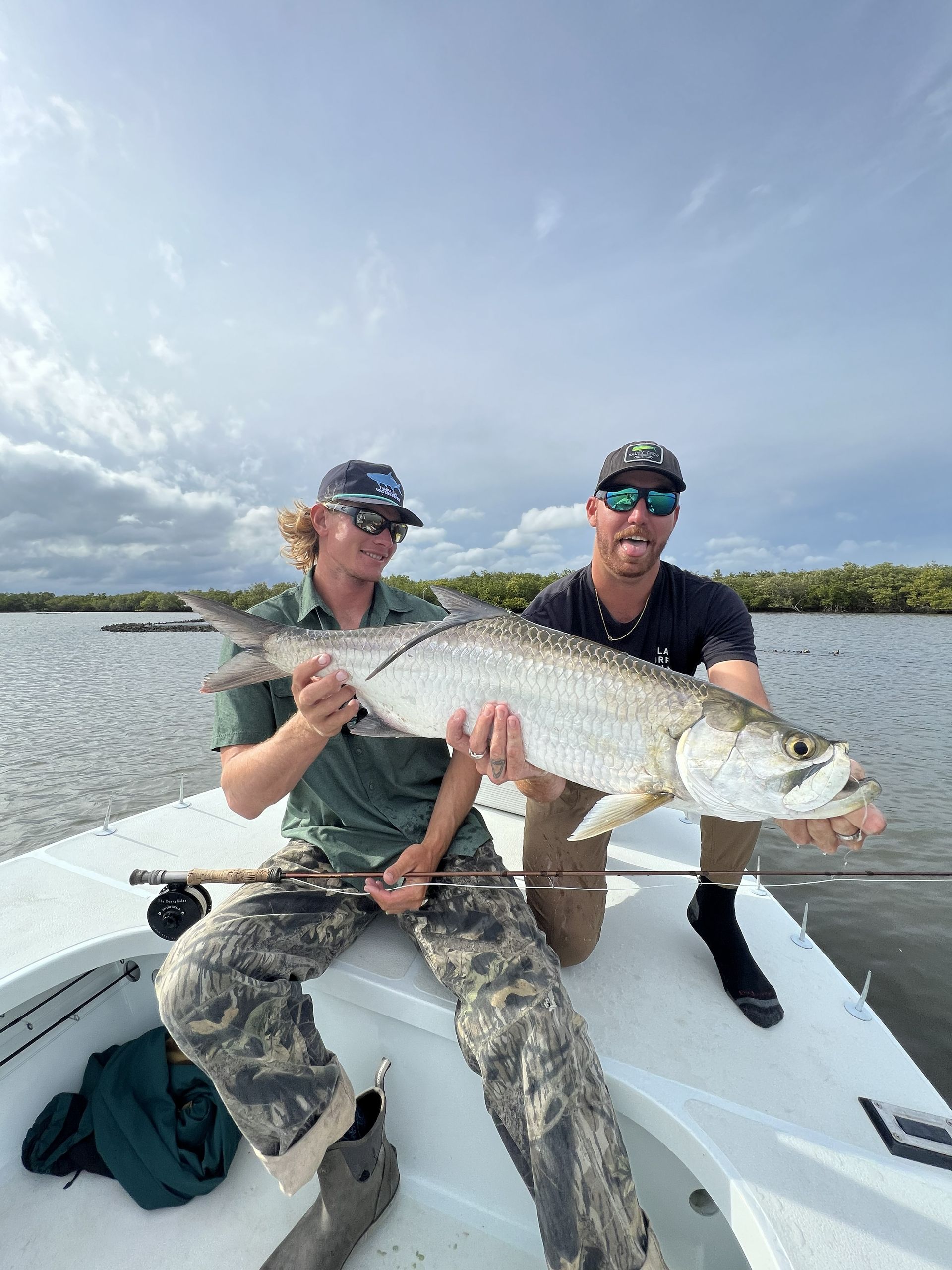 Two people on a boat holding a large fish under a bright sky on calm water.