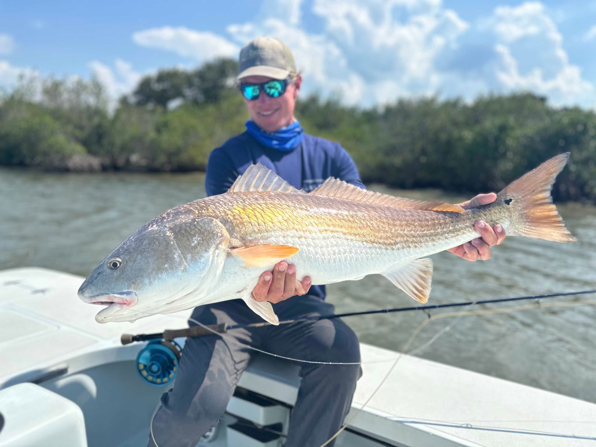 Angler holding a large redfish on a boat in shallow water, wearing sunglasses and a cap.