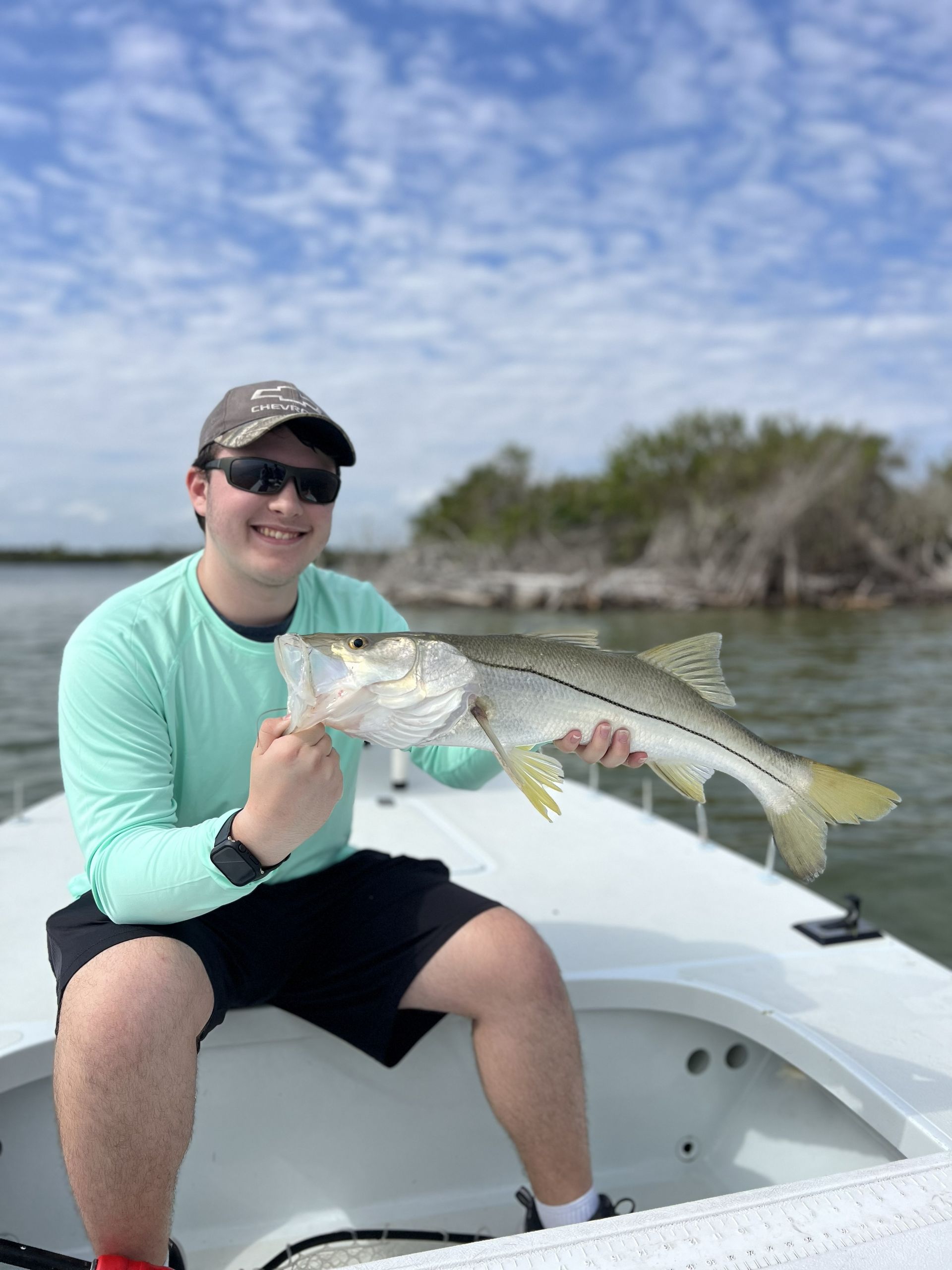 Person on a boat holding a large fish on a sunny lake under a partly cloudy sky.