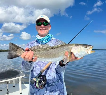 Smiling angler holding a large speckled fish on a boat under a blue sky