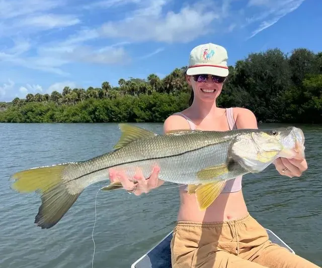 Person holding a large fish on a boat in a lake, wearing a hat and smiling under a blue sky.
