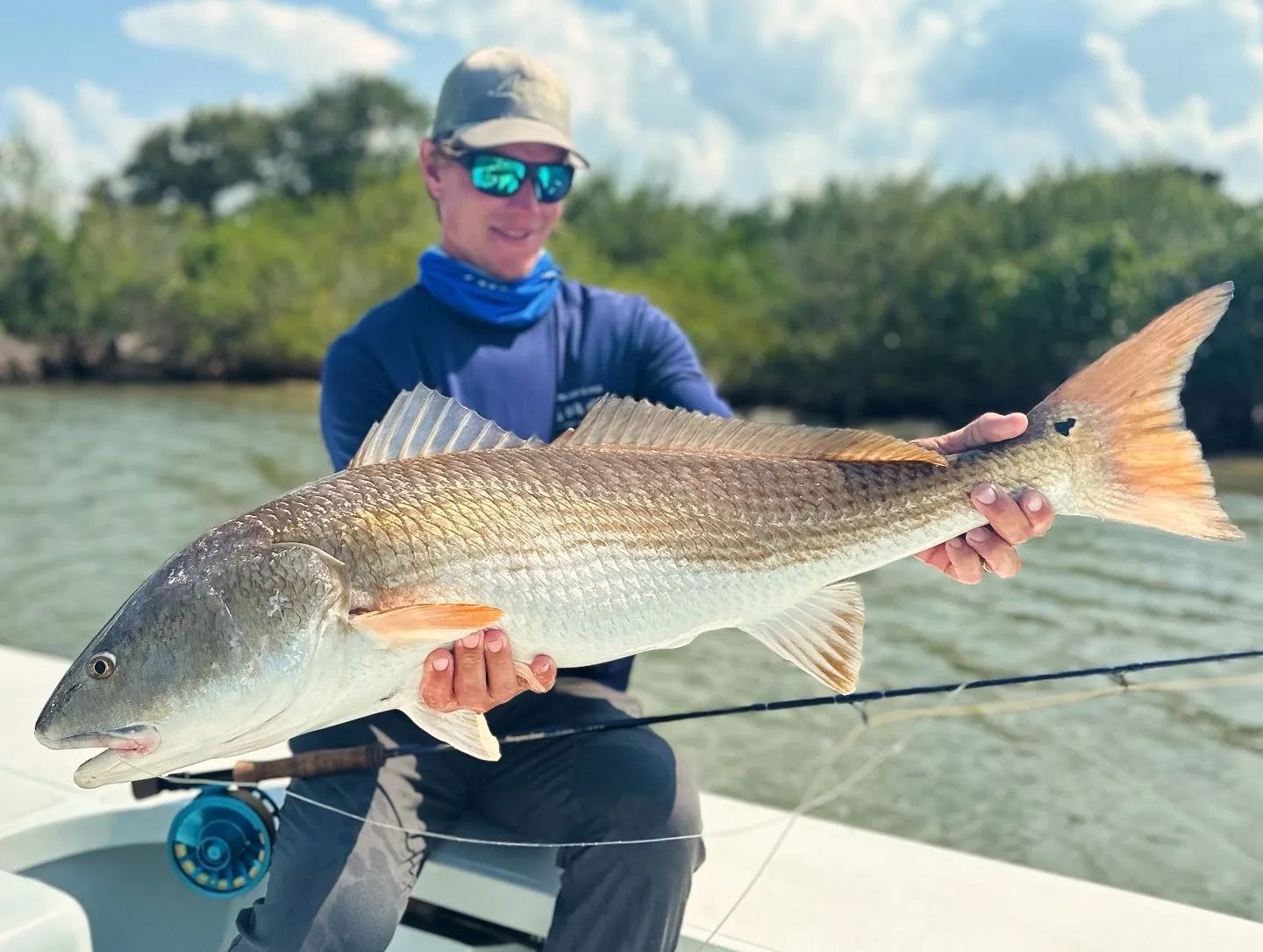 Angler holding a large silver fish on a boat in calm water, wearing blue shirt and cap.