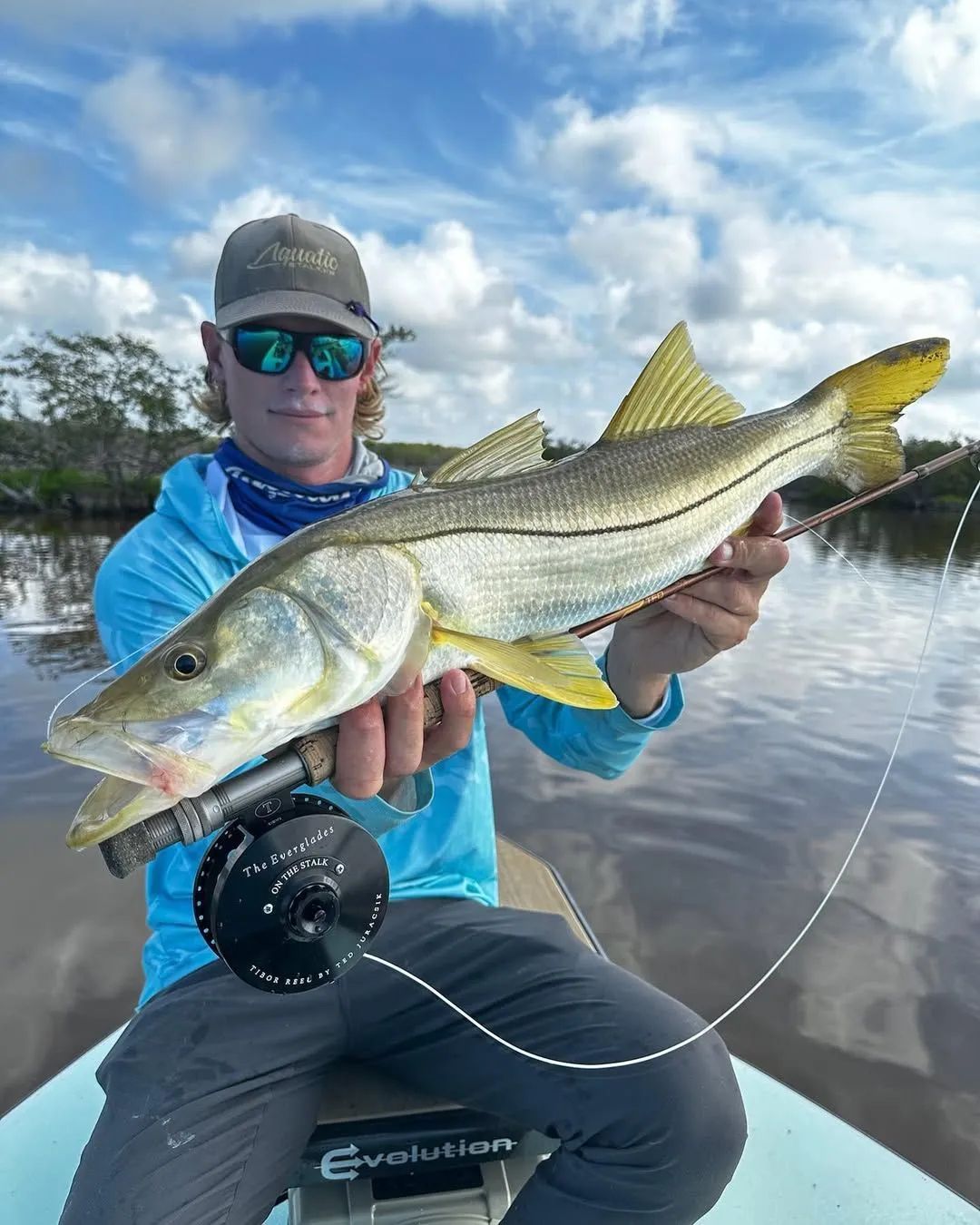 Angler holding a large striped fish on a boat, with water and cloudy sky behind.