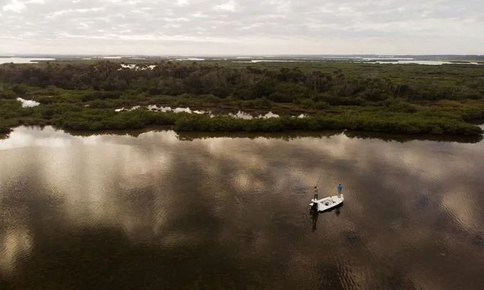 A small boat floats on calm brown water beside a mangrove forest under a cloudy sky.