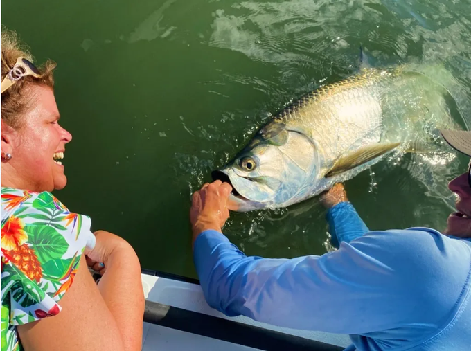 Two people on a boat holding a large fish at the water’s surface.