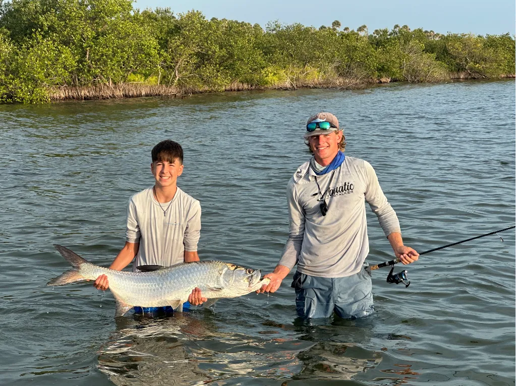 Two anglers in shallow water holding a large fish near a mangrove shoreline