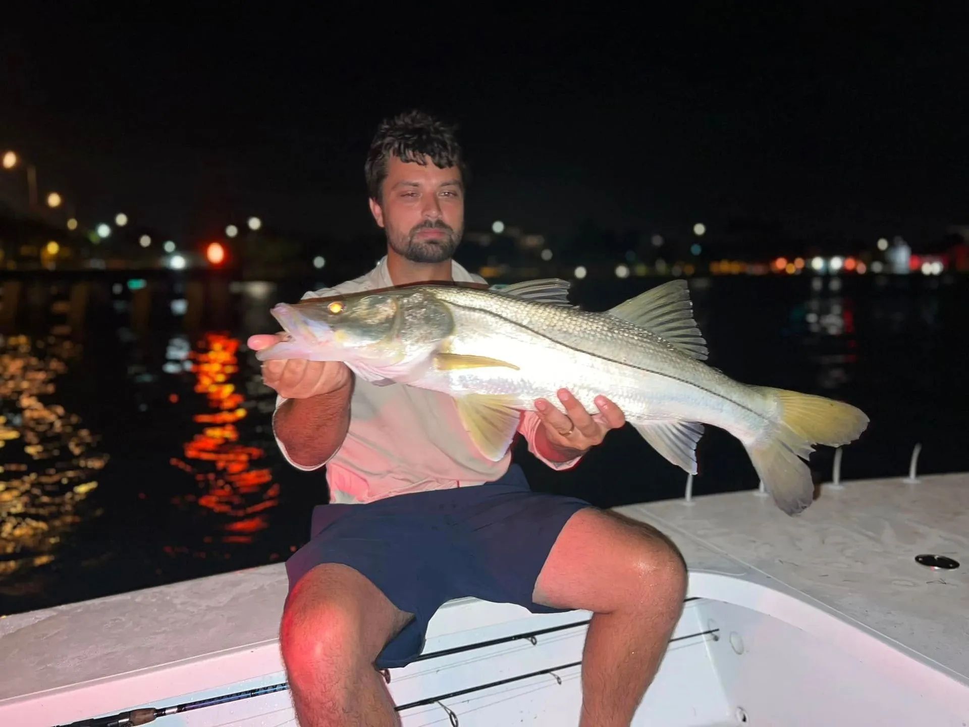 Man on a boat at night holding a large striped fish with city lights reflected on the water