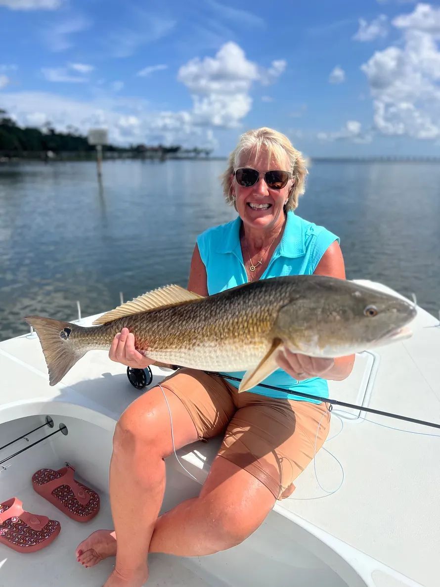 Woman smiling on a boat, holding a large fish on a sunny calm-water day.