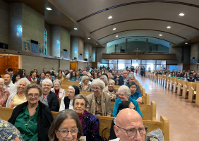Congregation seated in a church hall, facing the front with pews and arched ceiling.