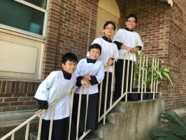 Four people in white shirts lean on a stair railing outside a brick building.