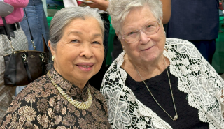 Two smiling women posing together indoors, one in black lace and one in white lace top.