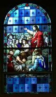 Stained-glass scene of a red-robed figure teaching a seated crowd beneath a blue arched window
