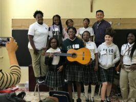 Group of students posing in a classroom, with one holding a guitar and a person taking the photo.