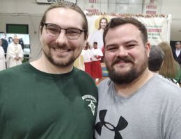 Two smiling men posing indoors at a crowded event, wearing green and gray shirts.