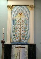 Altar with a painted floral tapestry, gold columns, and a cross above a white cloth