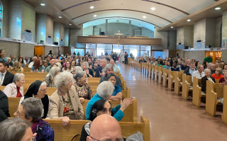 Crowded church hall with people seated in pews, facing a stage at the far end.