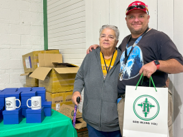Two people standing beside mugs and boxes, one holding a gift bag at a table in an indoor hall