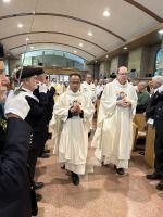 Priests in white vestments walking through a church aisle, flanked by ushers and applauding attendees