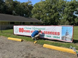 Person kneeling by a banner for 80 years of services at a roadside sign on a sunny day