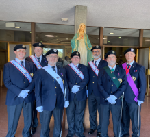 Seven men in formal attire stand indoors beneath a statue, wearing sashes and berets at a ceremonial event.