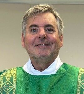 Smiling cleric wearing a green liturgical vestment and white collar indoors