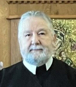 Portrait of a bearded man in a black clerical robe against a wood-paneled background with artwork